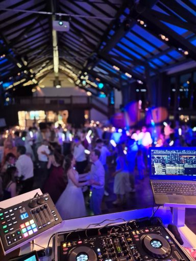 Gedämpfte Beleuchtung in einem Festsaal mit tanzenden Gästen und DJ-Pult im Vordergrund.